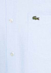 Lichtblauwe overhemd met knopen en een zak met een geborduurd groen krokodillogo. De stof heeft een gestructureerde weving en een gladde afwerking.