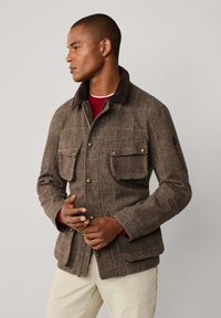 Bruine jasje met ruitpatroon en corduroy kraag, twee borstzakken en een getailleerde pasvorm. Draagt over een rode blouse, gemaakt van gestructureerde stof.