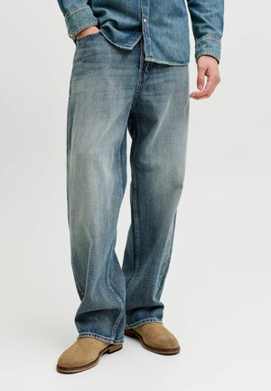 Uomo che indossa jeans azzurri a vestibilità comoda, una camicia di jeans e scarpe in camoscio color tan, in piedi con una mano in tasca.