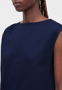 Gros plan d'une personne aux cheveux tressés portant un haut sans manches bleu marine avec un col rond simple.