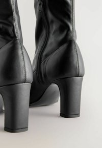 Črne usnjene škornje na peto imajo eleganten dizajn, gladko teksturo in detajl stranskega zadrge. Pete so čvrste in masivne.