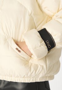 Giacca puffer di colore crema chiaro con finitura lucida, caratterizzata da un polsino nero a costine e un piccolo dettaglio logo metallico sulla manica.