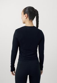 Top aderente nero a maniche lunghe realizzato in tessuto morbido con scollatura rotonda. Presenta un logo discreto sul retro, abbinato a leggins neri.