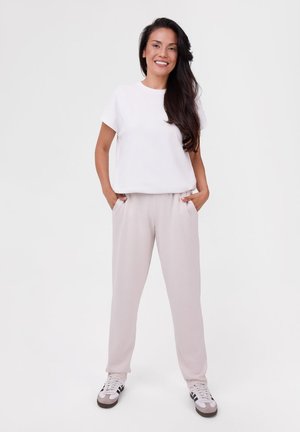 Frau steht mit den Händen in den Taschen, trägt ein weißes T-Shirt, hellbeige Hose und weiße Sneakers mit schwarzen Streifen.