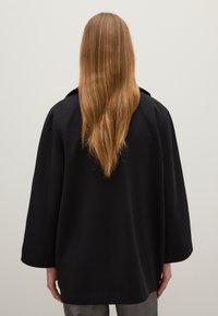 Schwarzer, übergroßer Mantel mit lockerer Passform und weitem Ärmel; ausgestattet mit einem klassischen Kragen und strukturiertem Stoff. Die Rückansicht zeigt lange, glatte Haare.