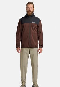 Bruin en zwart vest met rits en capuchon, gemaakt van zachte stof, gedragen over lichtbeige broek. Voorzien van een logo op de borst.