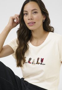 T-shirt crème avec des illustrations colorées de bottes de cowboy, présentant divers designs et motifs. Manches courtes et col rond.