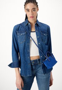 Camicia di denim di blu medio con maniche lunghe e bottoni a pressione, abbinata a un crop top bianco e a una mini borsa blu, con una texture liscia.
