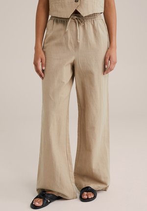 Pantalon large beige en tissu texturé avec une taille élastique et un cordon de serrage. Associé à des sandales noires avec un design de bride torsadée.
