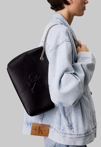 Musta nahkainen käsilaukku, jossa on kohopainettu logo ja hopeaketjuinen olkahihna. Malli pukeutunut vaaleaan farkkutakkiin.