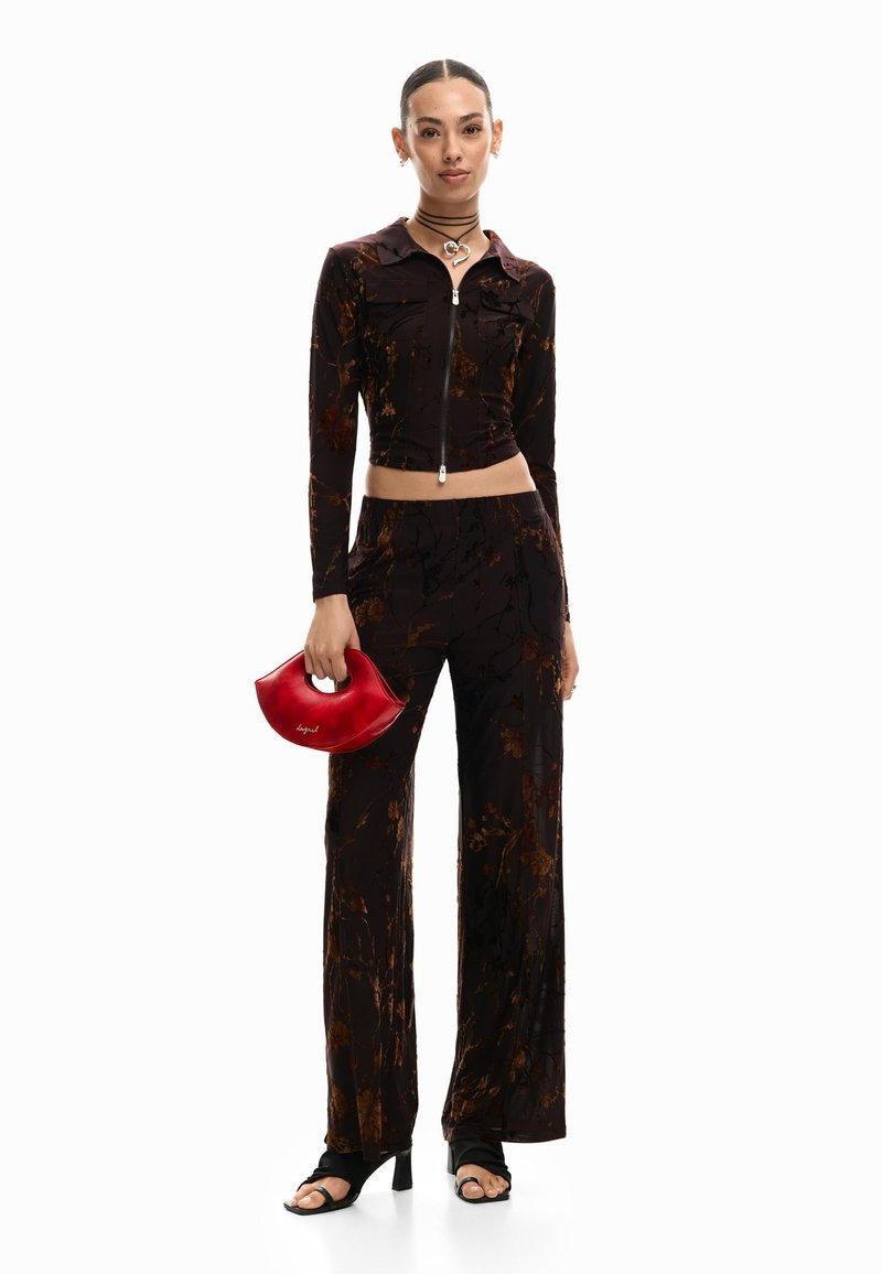 Jeune femme portant un crop top à fleurs foncées avec fermeture éclair et un pantalon large assorti, tenant un petit sac à main rouge et chaussant des talons noirs.
