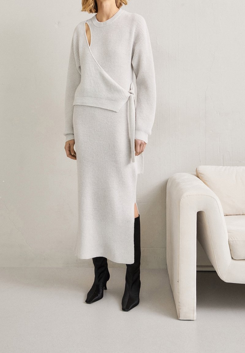 Ensemble asymétrique en deux pièces en tricot texturé gris clair avec un détail de fente sur le côté ; associé à des bottes noires jusqu'au genou.