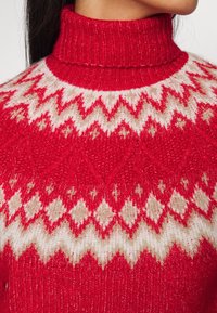 Red knitted sweater with a high ribbed collar. Features white and beige geometric patterns throughout. Soft texture with a cozy appearance.