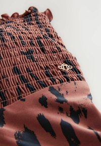 Rot-braunes Kleidungsstück mit geraffter Textur, das ein dunkles blaues abstraktes Muster sowie goldfarbene Hardware-Akzente aufweist. Weicher Stoff, geraffte Kanten.