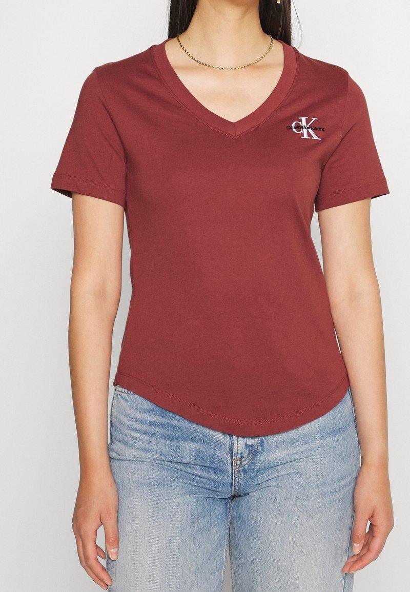 Donna che indossa una t-shirt Calvin Klein a maniche corte color bordeaux con scollo a V abbinata a jeans azzurri e una collana a catena dorata.