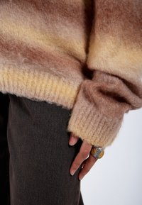Stickad tröja med mjuka gradientränder i ljusbrunt, gult och krämfärgat. Ribbad nederkant och ärmslut. Hand vilande på mörkbrunt tyg, bärande en silvring.