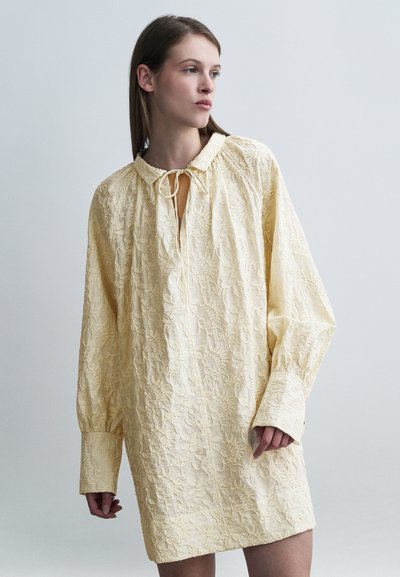 Robe jaune clair brodée avec un col, des manches longues et un détail noué au niveau de l'encolure. Elle présente un motif floral texturé.