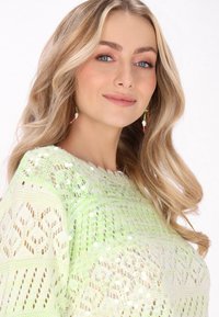 Strickpullover in Hellgrün mit Paillettenakzenten, der ein strukturiertes Muster und ein offenes Webdesign aufweist. Modell mit langen, welligen Haaren gestylt.