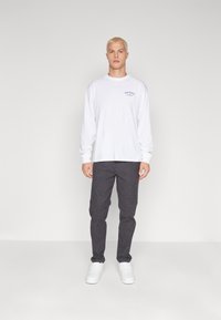 Maglietta bianca a maniche lunghe con logo "Dickies" e pantaloni grigi texturizzati, abbinati a sneakers bianche; il modello è in piedi davanti a uno sfondo neutro.