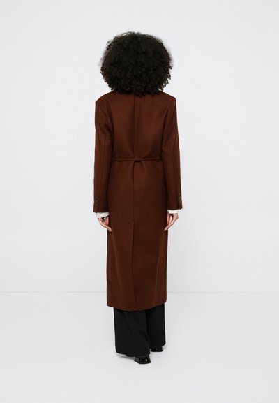 Ženska s kodrastimi lasmi, oblečena v dolg rjav plašč čez črne hlače, stoji z obrazom obrnjenim stran proti enobarvnemu belemu ozadju.