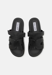 Crne natikače s ravnim potplatom i trakama od sintetičke kože s izrezbarenim uzorcima. Oznaka glasi "STEVE MADDEN."