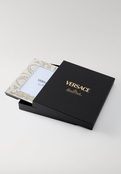 Melna kaste ar zeltu "Versace" logotipu, kas satur dekoratīvu rāmi krēmkrāsā ar smalku rakstu detaļām, atklājot tīru baltu ievietojumu.