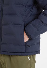 Navy puffer jacket with quilted design, featuring a side pocket and elastic cuffs for a snug fit. Lightweight fabric with a soft texture.