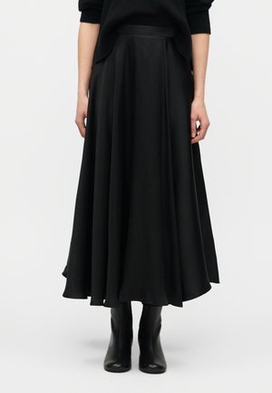 Γυναίκα που φοράει μια αέρινη μαύρη midi φούστα σε συνδυασμό με μαύρα μποτάκια αστραγάλου, στέκεται μπροστά σε ένα απλό ανοιχτό φόντο.