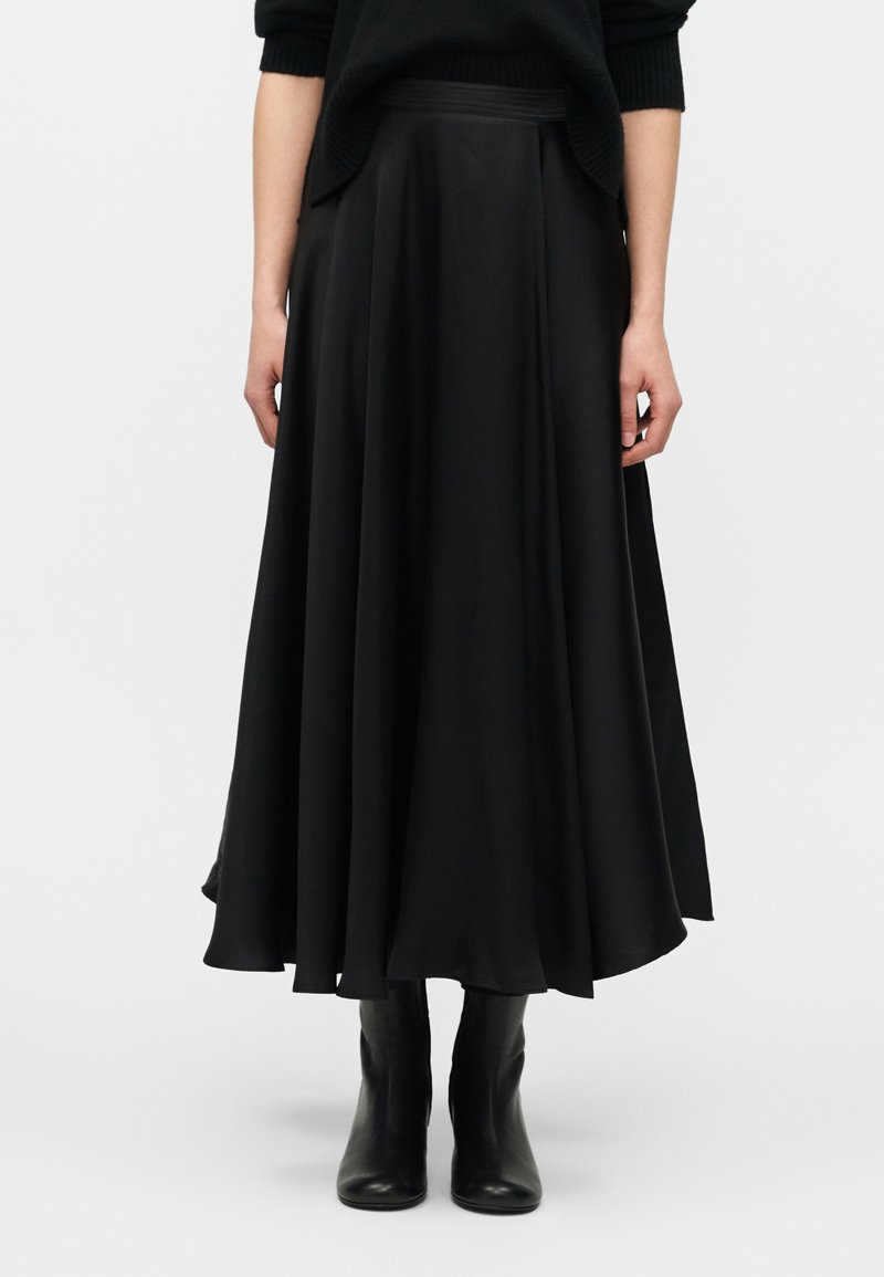 Γυναίκα που φοράει μια αέρινη μαύρη midi φούστα σε συνδυασμό με μαύρα μποτάκια αστραγάλου, στέκεται μπροστά σε ένα απλό ανοιχτό φόντο.