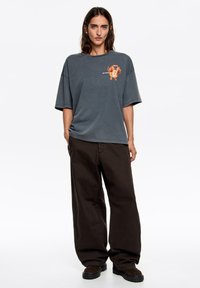Szara, oversize koszulka z grafiką psa na piersi, w połączeniu z luźnymi ciemnobrązowymi spodniami i czarnym obuwiem. Prosty wzór, swobodny styl.