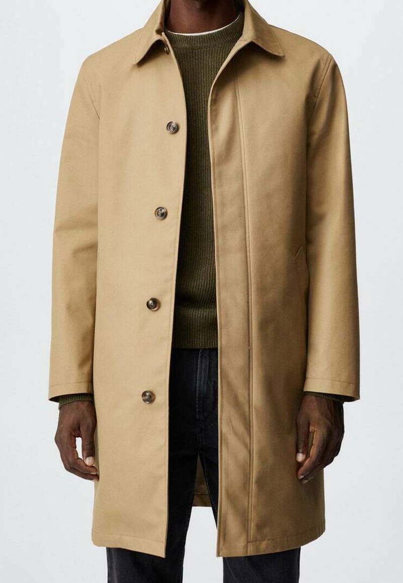 Beige, knielange jas met een uitgespreide kraag, sluiting aan de voorkant met knopen en twee zijzakken. Glad stoftextuur met een enkele plooi aan de achterkant.