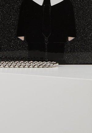 Pochette noire pailletée avec un design de smoking et une chaîne argentée posée devant sur une surface blanche.