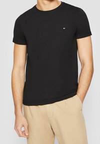 T-shirt noir en coton à col rond, avec des manches courtes et un petit logo rouge, blanc et bleu sur la poitrine gauche, associé à un pantalon beige.