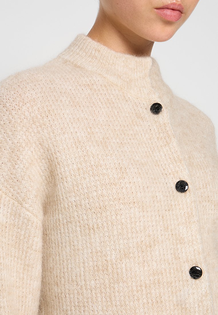 Beige gestrickte Strickjacke mit hohem Kragen, ausgestattet mit einem strukturierten Muster und drei schwarzen Knöpfen auf der Vorderseite. Weiches, gemütliches Material.