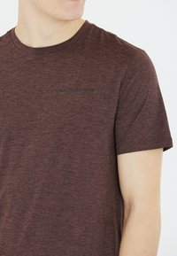 Ruskea heather T-paita pyöreällä pääntiellä ja lyhyillä hihoilla; rinnassa on herkkä musta "ENDURANCE" -logo. Pehmeä kankaan rakenne.