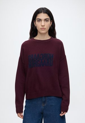 Jeune femme aux longs cheveux foncés porte un pull ample bordeaux avec un texte bleu miroir et un jean bleu, debout devant un fond blanc uni.