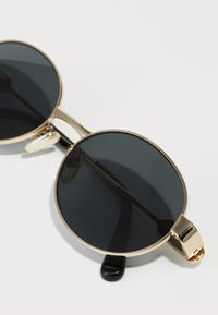 Gafas de sol redondas con lentes negros, montura de metal dorado y un puente nasal prominente. Presentan puntas de patilla delgadas en negro y bisagras de resorte.