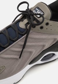 Grå atletisk sko med mesh-textur, svarta detaljer och en vit sula. Har en märkesetikett och korslagda snören.