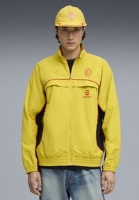 Yellow lightweight jacket with red accents, zip closure, and logo on chest. Black side panels and matching cap complete the sporty look.