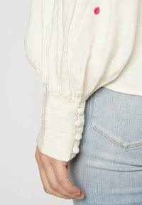 Vit blus med en texturerad ärm som har veck och klädda knappar. Bärs över ljusblå denimjeans.