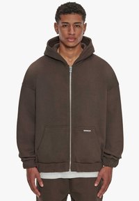 Hoodie zippé marron avec une grande capuche, poches avant et poignets côtelés. Fabriqué en tissu doux, avec une coupe décontractée et un branding minimal.