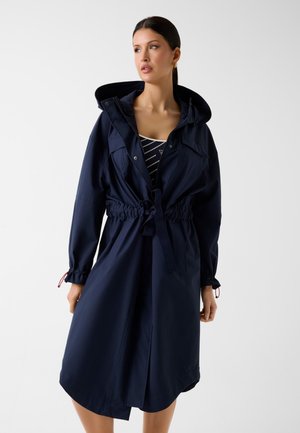 Kobieta ubrana w granatowy płaszcz z kapturem, z wcięciem w talii, elastycznymi mankietami i pasiasty top pod spodem, stojąca na jednolitym białym tle.