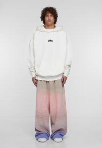 Felpa bianca con tasca frontale e logo nero, abbinata a pantaloni larghi con un motivo ombre che va dal rosa al viola.