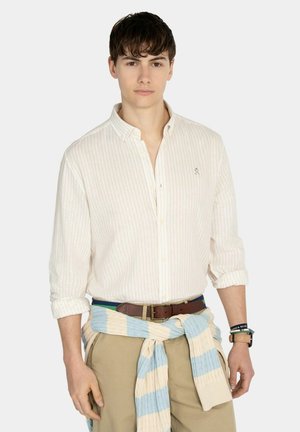 Joven con una camisa clara de rayas con botones, pantalones beige, cinturón marrón, jersey azul y crema atado alrededor de la cintura y pulseras.