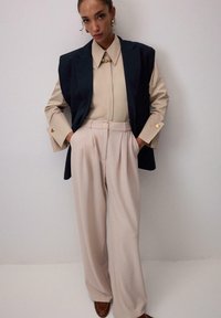 Béžová košeľa s golierom, tmavá vesta, béžové plisované nohavice, zlaté gombíky ako akcenty a hnedé topánky; hladká tkanina a prispôsobený strih.