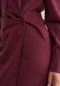 Artimųja nuotrauka, kurioje matyti ranka, padėta šalia asmens, apsirengusio prigludusia tamsiai burgundiška suknele su sagčių detalėmis ties juosmeniu.