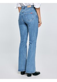 Žena nosící světle modré rozšířené džíny a černé kotníkové boty na podpatku, zobrazená zezadu na jednobarevném světlém pozadí.