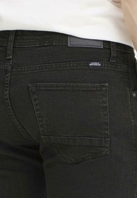 Jeans negri din denim cu un buzunar pe spate și o textură netedă. Prezintă o etichetă gri la talie cu text. Designul este simplu și slim.