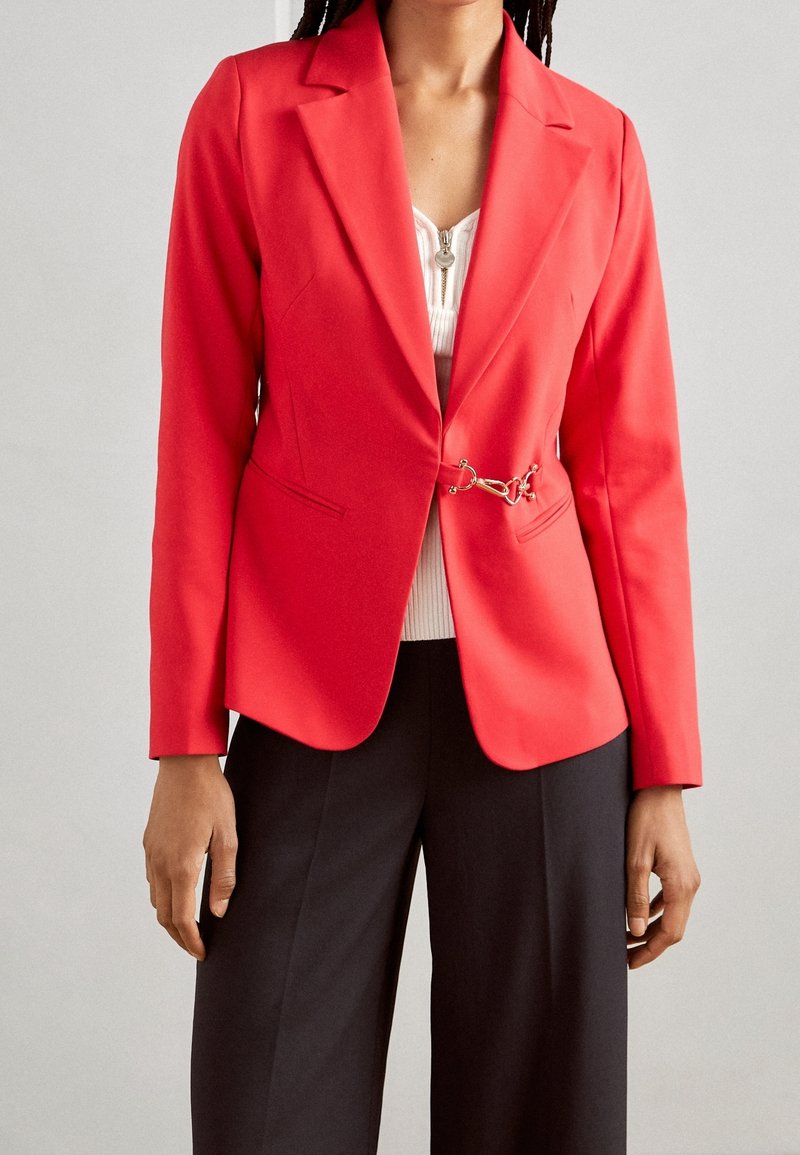 Rød blazer med skræddersyet pasform, blødt materiale, notch-lapel og kædeaccent i taljen. Båret over en hvid ribbet top og sorte bukser.