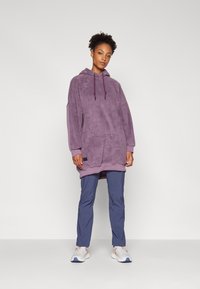 Lilla fleece oversized hettegenser med en frontlomme og snøring. Kombinert med blå tapered bukser og hvite joggesko med oransje detaljer.